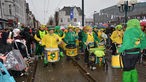 Zu sehen ist ein Karnevalsverein, die trommelnd und mit grüngelben Kostümen durch die Straße zieht.