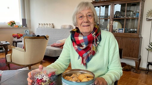 Die ältere Frau sitzt an einem Holztisch mit einer offen Keksdose, mit Keksen gefüllt.  