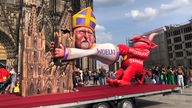 Die Großskulptur des Düsseldorfer Wagenbauers Jacques Tilly zeigt Kardinal Woelki, der sich - vom Teufel namens "Missbrauchsskandal" bedroht - verzweifelt an den Kölner Dom klammert und dadurch einen seiner Türme zum Einsturz bringt.