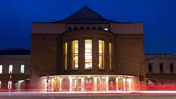 Auf dem Foto ist das Wuppertaler Operngebäude im Dunkeln: Ein beleuchtetes Betongebäude mit sehr großen Fenstern und einem ausladenden Foyer.