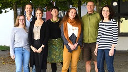 Gruppenbild mit Erwachsenen vor weißer Hauswand und grünen Bäumen