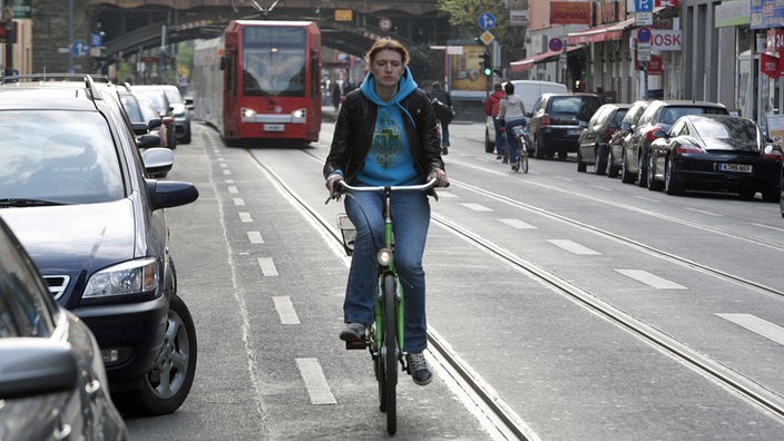 Eine Frau fährt mit dem Rad die Zülpicher Straße entlang, am Straßenrand parken Autos, im Hintergrund eine KVB-Bahn