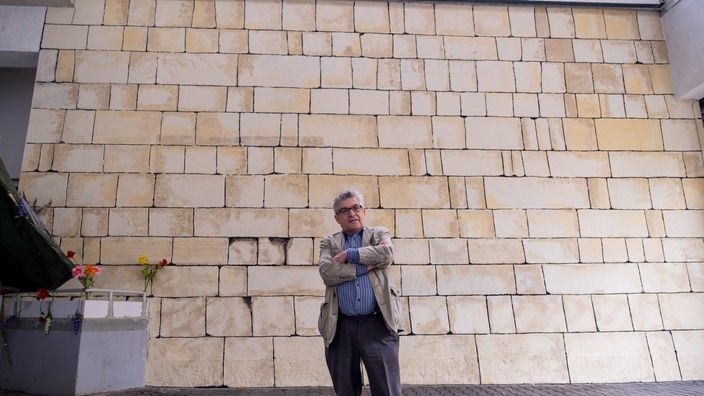 Ein Mann steht vor eine großen hellen Mauer.