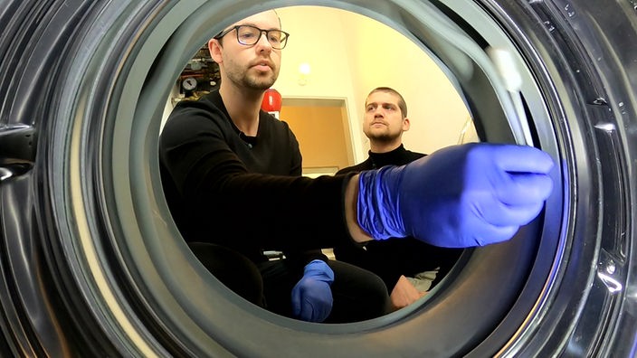 Der Forscher entimmt aus einer Wäschetrommel mit einem riesigen Wattestäbchen und blauen Handschuhen eine Probe