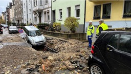 Ein Auto neigt mit der Vorderseite Richtung des Straßenrisses, der sich aufgrund des Wasserrohrbruchs geöffnet hat. Auf der Staße stehen zwei Männer mit Warnwesten.