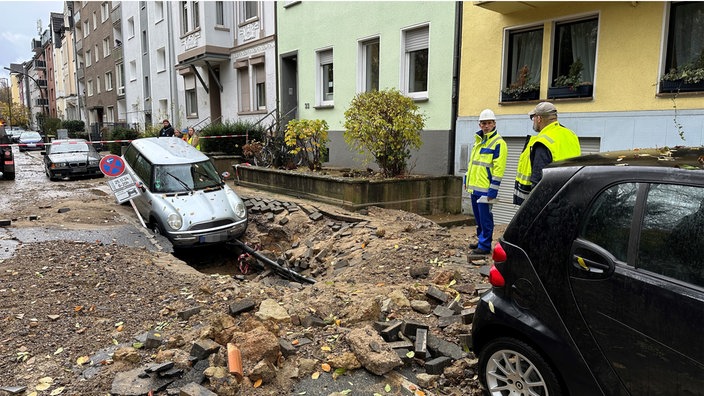 Ein Auto neigt mit der Vorderseite Richtung des Straßenrisses, der sich aufgrund des Wasserrohrbruchs geöffnet hat. Auf der Staße stehen zwei Männer mit Warnwesten.