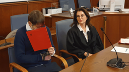 Beschuldigter Mann aus Hamm verdeckt mit Mappe sein Gesicht, rechts daneben sitzt seine Anwältin zum Verfahrensauftakt