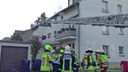 Eine Gruppe Feuerwehrleute steht vor einem Haus, von rechts ins Bild erstreckt sich eine Drehleiter mit einem löschenden Feuerwehrmann im Korb. Man sieht ein verrauchtes Fenster am Haus, die Wand ist unterhalb der Dachkante verrußt.