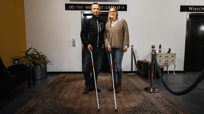Jane und Jürgen stehen vor einem Schild mit der Aufschrift Stockfinster und halten einen Blindenstock.