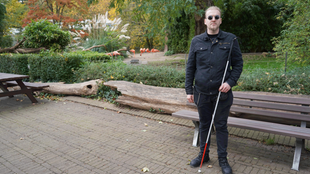 Ein dunkel gekleideter Mann mit Sonnenbrille und Blindenstock steht vor einem Tiergehege im Zoo