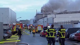Meherere Feuerwehrfahrzeuge und Einsatzkräfte stehen auf einer Straße in einem Gewerbegebiet in Holzwickede. Im Hintergrund steigt dunkler Rauch auf.