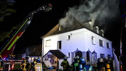 Rauch steigt aus dem oberen Geschoss des Mehrfamilienhauses in Gevelsberg. Davor stehen Einsatzkräfte und ein Feuerwehrfahrzeug mit Drehleiter.