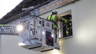 Auf dem Foto ist ein Feuerwehrmann in einem Korb einer Drehleiter vor einem Fenster. Darüber ist ein abgebranntes Dach.