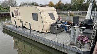 Christian Gröll aus Essen sitzt auf seinem Boot, es ist ein Katamaran mit einer Plattform und einem alten Wohnwagen ohne Räder darauf