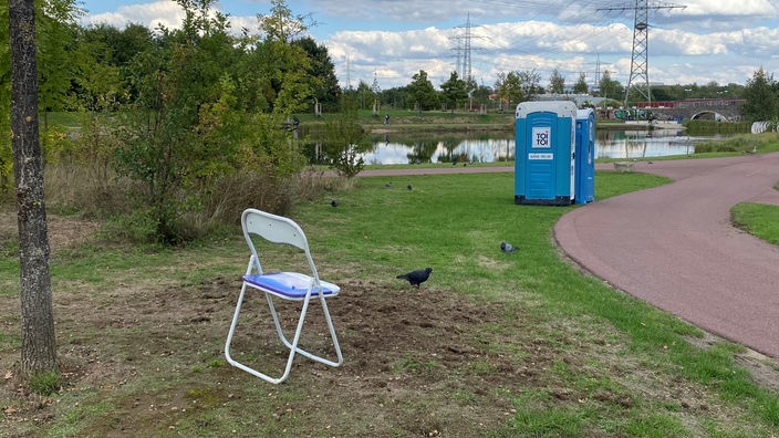 In einem Park steht ein Stuhl, weiter hinten im Bild sieht man zwei Dixie-Klos auf dem Rasen stehen.