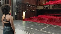 Auf dem Foto ist eine Frau mit dunkler Hautfarbe, die von der Bühne aus auf leere Publikumsplätze im Dortmunder Theater schaut.