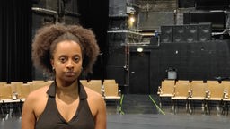 Auf dem Foto ist eine Frau mit dunkler Haut, die auf einer Theaterbühne steht und mit entschlossenem Blick nach vorne schaut.