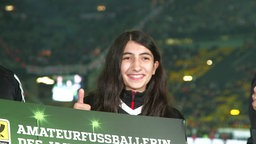 Auf dem Foto ist eine junge Frau mit dunklen Haaren, die lächelnd hinter einem Schild steht. 