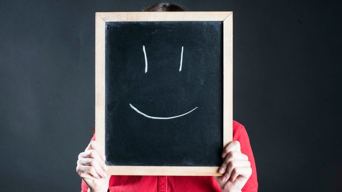 Eine Frau hält eine Tafel vor ihr Gesicht, auf die mit Kreide ein Smiley gemalt ist