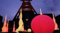 Das Bild zeigt den Förderturm der Zeche Zollverein bei Nacht, davor sind gelb und rot leuchtende Bälle und Objekte, die wie züngelnde Flammen aussehen, zu sehen.