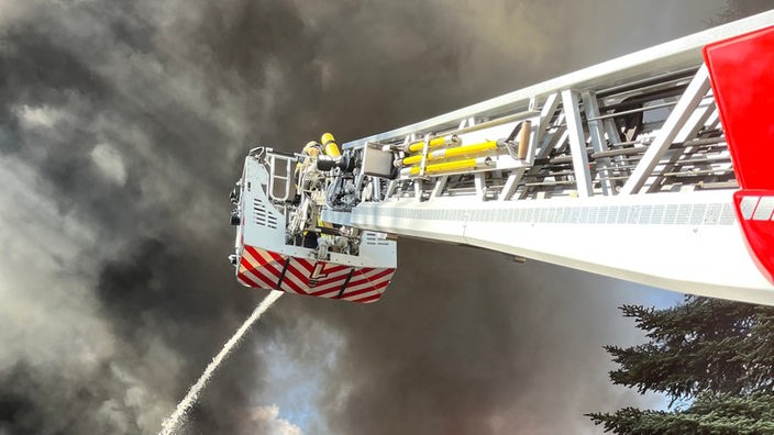Feuerwehrleute stehen weit oben auf einer Drehleiter und versuchen den Brand zu löschen. Im Hintergrund sind dicke Rauchwolken zu sehen.