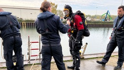 Menschen stehen am Wasser. In der Mitte steht ein voll ausgerüsteter Taucher.