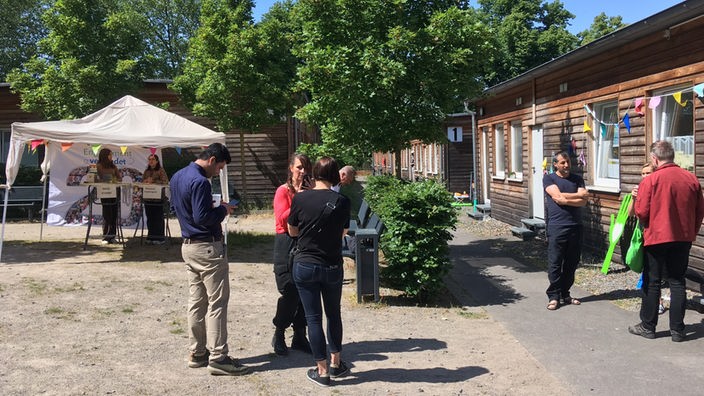 Mehrere Menschen stehen vor einem Info-Pavillon, im Hintergrund Holzhäuser