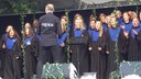 Ein Chor steht auf einer Bühne, die Chormitglieder sind einheitlich in blau und schwarz gekleidet, auf dem Oberteil des Chorleiters steht "rejoice"