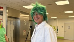 Ein Mann mit weißem Kittel und einer grünen Perücke steht in einem Krankenhausflur und lacht. 