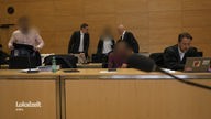 Zu sehen sind sechs Menschen, drei davon im Anzug und bei dreien sind die Gesichter verpixelt. Sie befinden sich in einem Gerichtssaal.