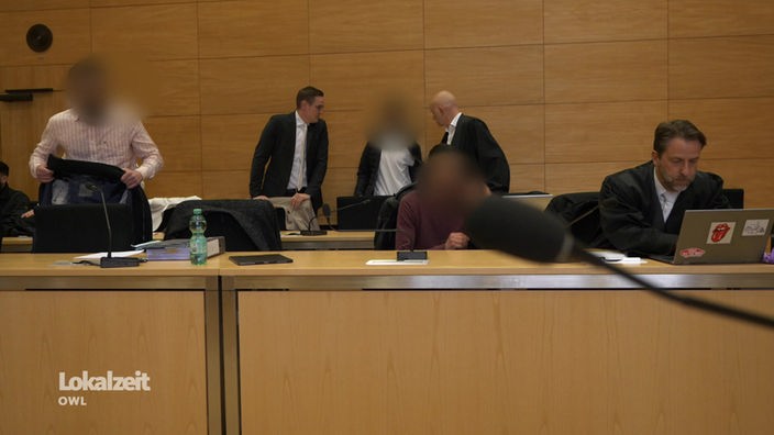 Zu sehen sind sechs Menschen, drei davon im Anzug und bei dreien sind die Gesichter verpixelt. Sie befinden sich in einem Gerichtssaal.