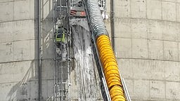 Auf dem Foto ist ein Feuerwehrmann, der durch einen Schwall aus Schaum über eine Leiter steigt. Daneben ist ein großer Schlauch, der Schaum gegen das Silo pumpt.