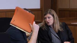 Auf dem Foto ist ein Mann, der sich eine organgene Mappe vor sein Gesicht hält. Eine blonde Frau, seine Anwältin, redet mit ihm.