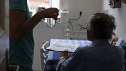 Ein älterer Mann sitzt an einem Tisch. Eine weitere Person reicht ihm ein Glas Wasser.