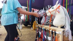 An einem Stand auf dem Hobby Horsing Turnier hängt Zubehör für Steckenpferde. Ein kleines Mädchen schaut es sich an.