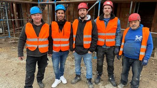 Mitarbeiter des Gerüstbauunternehmens mit Warnwesten und Helmen, im Hintergrund eine Baustelle