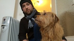Mieter Dennis Klee mit Mütze und Schal bei 12,1 Grad in seiner Wohnung. Neben ihm sein Hund, dahinter die Heizung