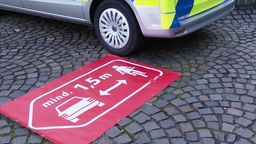 Neben einem Polizeiauto markiert ein roter Bereich auf dem Boden, wie groß der Abstand von 1,5 m sein muss