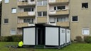 Das Bild zeigt einen Toiletten- und Duschcontainer vor einem Mehrfamilienhaus.