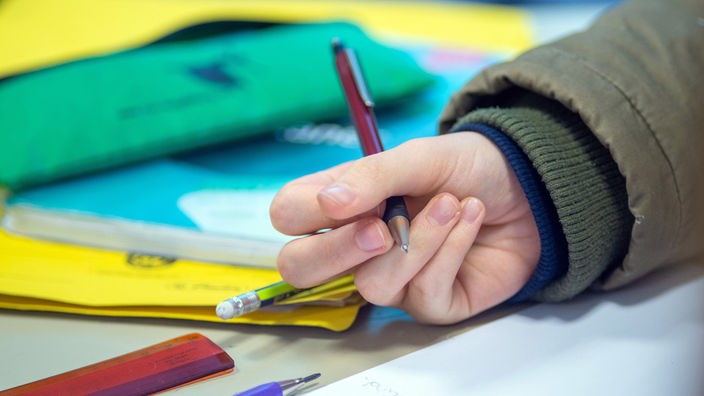 Auf dem Foto ist eine Kinderhand, die einen Stift hält. Daneben sind Hefte und Stifte.