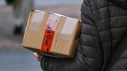 Symbolfoto: Eine Frau in schwarzer Jacke hat ein großes Paket im Arm