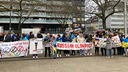 Das Bild zeigt Menschen, die in Essen gegen IOC-Präsident Thomas Bach protestieren. Sie haben sich in einer Reihe aufgestellt und haben Plakate in den Händen.