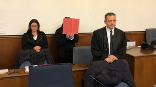 Der Angeklagte hält sich eine Mappe vor das Gesicht, daneben seine Anwälte