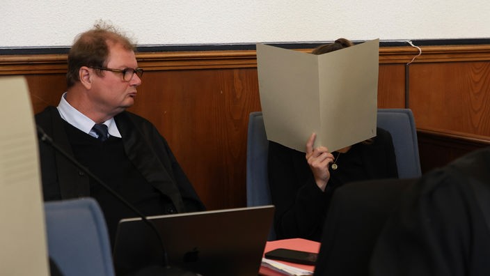 Frau schützt ihr Gesicht mit einer Akte. Neben ihr sitzt ihr Anwalt.