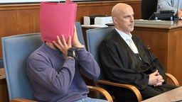 Angeklagter hält sich Akte vor das Gesicht, neben ihm sitz sein Anwalt
