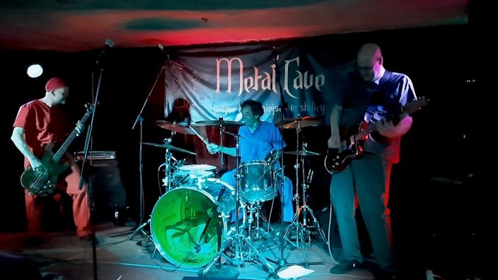 Man sieht einen dunklen Raum mit drei Personen, die vor einer Wand mit einem Banner stehen.  Der Mann links hat eine Bassgitarre in der Hand, der Mann in der Mitte spielt am Schlagzeug und rechts sieht man den Gitarristen.
