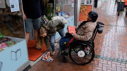 Eine Frau und ein Mann im Rollstuhl legen eine Rampe vor eine Stufe vor einem Buchladen.