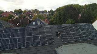 Auf dem Foto ist ein Hausdach mit blauen Ziegeln, es ist voller Solarzellen. Zwischen den Solarzellen schaut ein Mann aus einem Dachfenster.