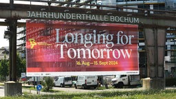 An der Jahrhunderthalle in Bochum hängt en großes Plakat für die Ruhrtriennale 2024 mit dem Titel "Longing for Tomorrow"