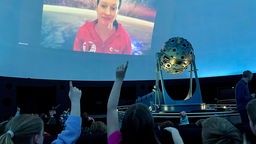 Kinder zeigen in die Kuppel des Planetariums, auf einer Videoleinwand sehen eine Astronauten-Kandidatin